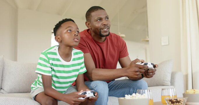 African American father and son grabbing white game controllers with TV coming alive on sofa
