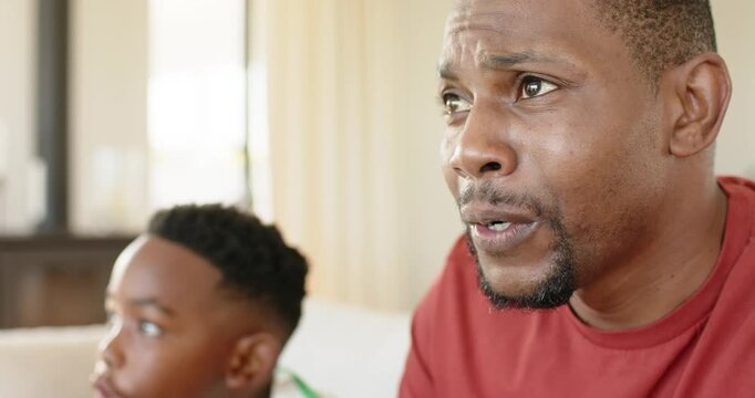 African American father and son reacting to off-screen TV program and watching on living room couch