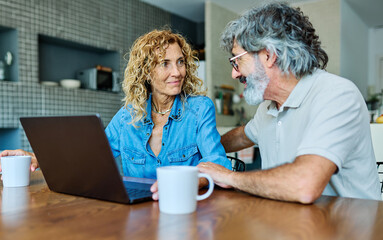 Portrait of a lovely senior mature couple using a laptop together and having fun drinking coffee or tea sitting at a table at home