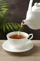 Pouring hot tea into cup from teapot at wooden table, closeup