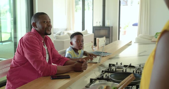 African American family father speaking with mother stirring pan, son pausing typing at laptop