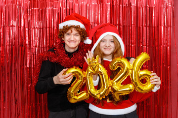Happy friends in a santa costume holding golden balloons with the shape 2026 on a red background.