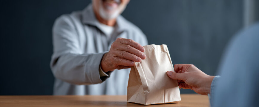 Close-up of elderly man receiving a brown paper bag from another person across a wooden table in a casual indoor setting - Powered by Adobe