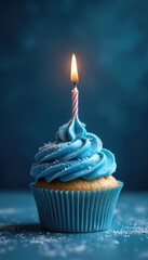 A single blue frosted cupcake with a lit candle sits on a blue surface. The dessert is decorated with white sprinkles for a celebratory occasion. It looks delicious and sweet.