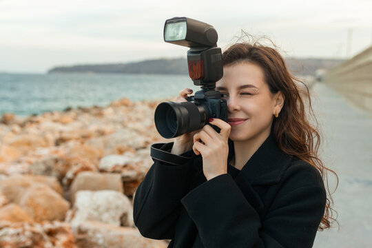Young female photographer taking pictures outdoors by the sea, smiling while holding a professional camera with flash, capturing moments on a rocky coastline at sunset.