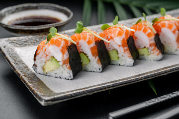 Sushi rolls with salmon and avocado sit on a dark plate. A small bowl of soy sauce rests beside them. Green leaves add to the setting
