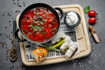 A bowl of red soup is placed on a wooden tray along with bread, sour cream, and fresh vegetables, prepared for a midday meal with family or friends