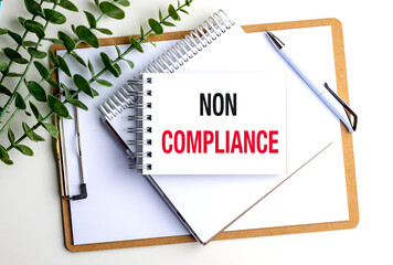 Non compliance concept shown on a notepad with office supplies and plant on white background
