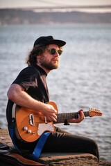 Hipster street musician in black playing electric guitar in the street sitting on pier embankment on sunset