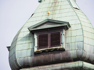 Close-up of a weathered copper-green turret or dome with a small louvered window under an overcast sky in the city. © Олексій Остистий
