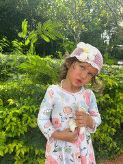 A small, possessive girl in a pink hat, holding flowers and a disgruntled expression, against a backdrop of tropical greenery. A three-year-old emotional girl.