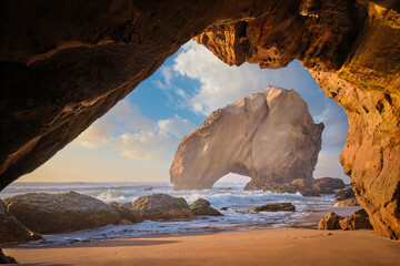 Penedo do Guincho, a large boulder rock arch at Praia da Santa Cruz, Portugal, with ocean waves and sandy beach as seen from grotto cave on sunset