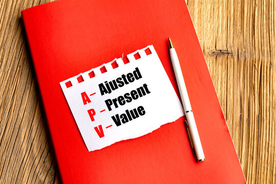Acronym APV adjusted present value written on paper with red notebook and pen on wooden desk