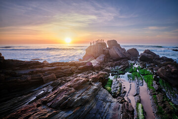 Scenic rock formation at Praia da Santa Cruz, Portugal, with ocean waves and vibrant sunset sky reflecting on the rocks.