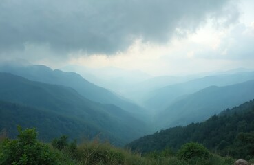 Naklejka premium View from mountain top shows many layered hills and green forest. Mist covers valleys, creating a hazy, blue landscape. Cloudy sky above peaceful, vast nature scene. Distant mountains fade in fog.