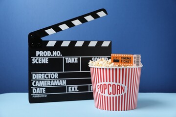 Tasty popcorn in paper cup, movie ticket and clapper on table against blue background