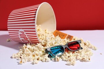 Tasty popcorn in paper cup, movie ticket and 3D glasses on white table against red background, closeup