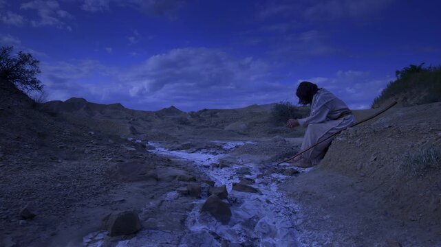 Jesus alone in a desolate landscape at night. Dressed in traditional white robes, the traveler appears to be resting with a wooden staff at his side, surrounded by cracked earth and distant hills.