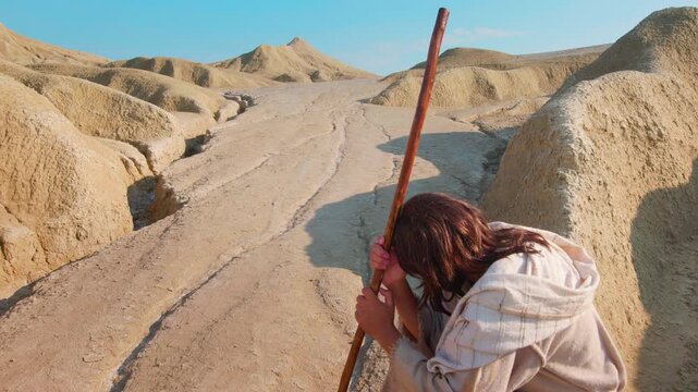 Revealing shot of Christ in a desolate landscape. Dressed in traditional white robes, the traveler appears to be resting with a wooden staff at his side, surrounded by cracked earth and distant hills.