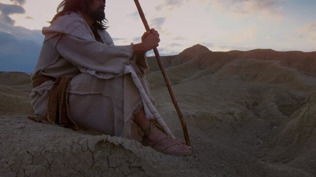 An epic scene at twilight depicting Jesus alone in a desolate and arid landscape. Dressed in traditional white robes, the traveler appears to be resting with a wooden staff at his side.
