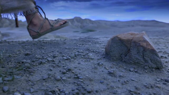 Footsteps in the wilderness at night. Slow motion footage of a detailed close-up showing the feet of a traveler clad in ancient rope sandals, standing beside a prominent rock on cracked earth.