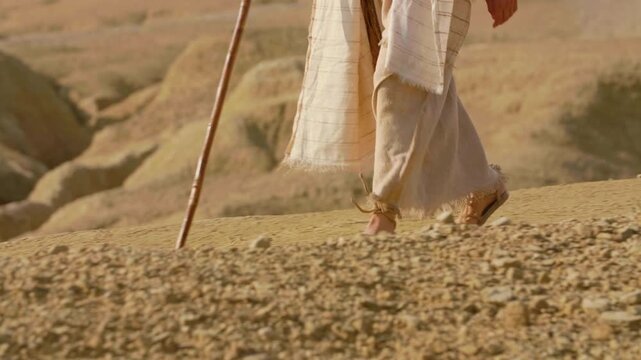 Slow motion close-up showing the feet of a traveler clad in ancient rope sandals, standing beside a prominent rock on parched, cracked earth. Christian and spiritual concept