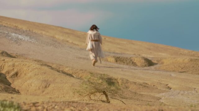 Back view of a robed wanderer crossing a rugged, sunlit desert terrain with natural clay hills and soft contours. Atmospheric wilderness scenery suggesting pilgrimage and spiritual journey.