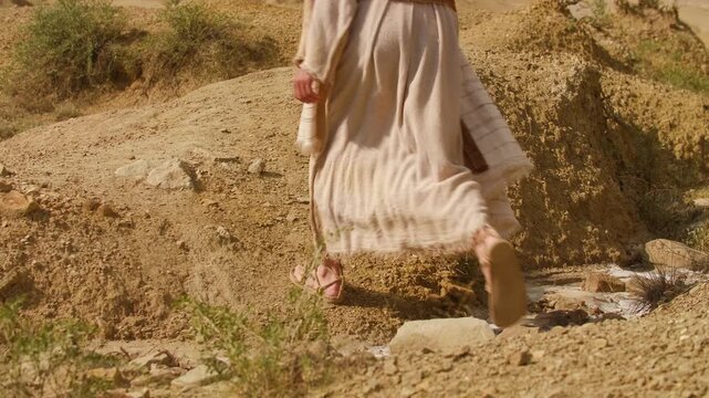 Revealing shot of a human representation of Christ in traditional biblical-style clothing, walking through an arid desert landscape, surrounded by sculpted sand formations and warm golden light.
