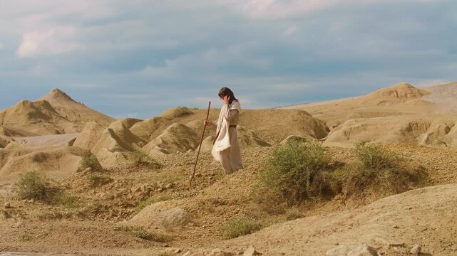 Depiction of Messiah in ancient-style clothing walks through a barren desert landscape. Cinematic scene evoking solitude, spirituality and timeless exploration. Christian concept. Slow motion footage