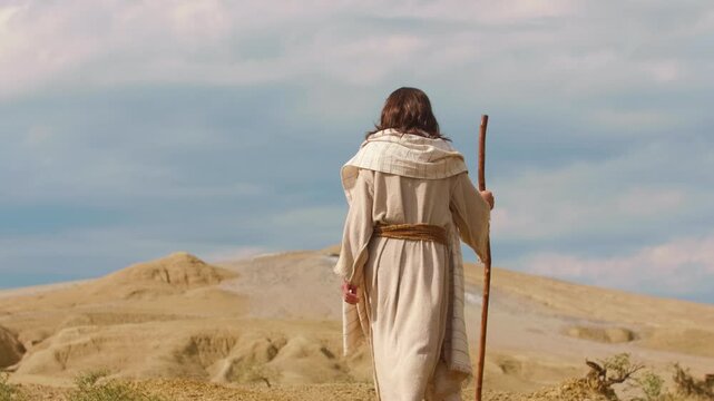 Solitary figure of Jesus in traditional clothing walking along a dry and eroded valley surrounded by dramatic desert mounds. Wild environment, symbolizing endurance, faith and personal quest.