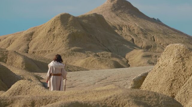 Representation of Christ, dressed in traditional white robes, explores an austere and sculptural desert environment. The natural formations evoke a sense of temptation or timeless wandering.