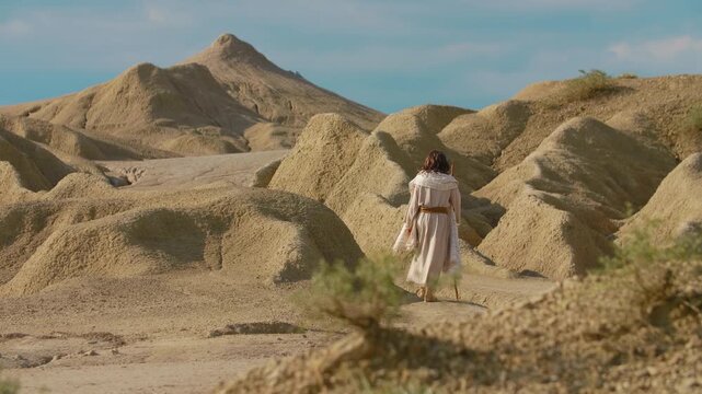 Wide Shot of a Traveler dressed in simple biblical-style robes exploring an austere and sculptural desert environment. The earthy tones of the natural formations evoke a sense of timeless wandering