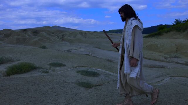 Representation of Christ, wearing ancient robe, walks away into the night, crossing a barren and rugged landscape. A dry white stream runs through the foreground, against a dramatic blue sky