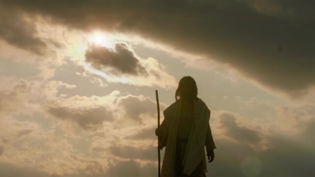 Alone Messiah in ancient robes standing atop a barren hill, arms raised wide in a gesture of deep prayer, adoration and spiritual triumph over the surrounding wilderness.