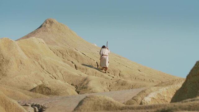 A robed figure walks through a sandy desert holding a wooden staff. Christian and spiritual concept. Slow motion Shot