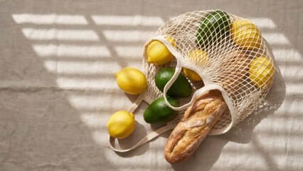 Fresh produce in a reusable mesh grocery bag on a linen surface. Sustainable shopping with lemons, avocados, and bread. Healthy eating flat lay with natural light and shadows