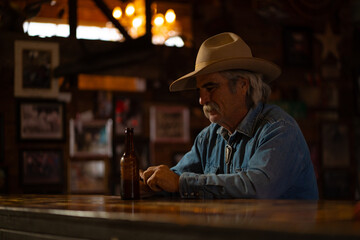 Man With Mustache Wearing Cowboy Hat and Denim Shirt Sitting and Drinking Beer at Bar in Rural Rustic Western Saloon Bar