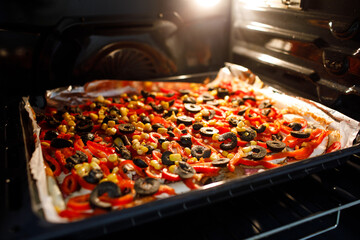 Extreme close-up of a homemade pizza topped with colorful vegetables, black olives, and corn, baking inside a glowing oven