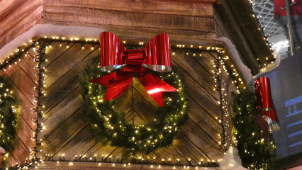 Colorful holiday wreath with large red bow and festive lights adorns a wooden structure in a winter setting