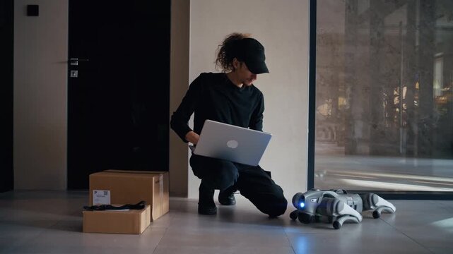 A technical delivery specialist activates an automated delivery system, using a laptop to configure a quadrupedal robot that will transport a cardboard package.