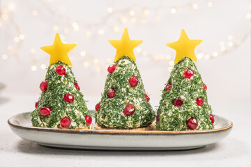 Three decorative cheese trees are placed on a table. Each tree is topped with a star and decorated with herbs and red fruits. Soft lights shine in the background, creating a holiday mood.