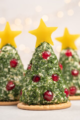 Three decorative cheese trees are placed on a table. Each tree is topped with a star and decorated with herbs and red fruits. Soft lights shine in the background, creating a holiday mood.