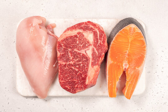Three pieces of raw meat are placed on a cutting board in a kitchen. The selection includes chicken breast, beef steak, and salmon fillet. The kitchen has a clean and simple look.