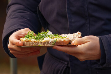 A person holding an open-faced sandwich topped with fresh herbs at the Naplavka farmers market in Prague, Czech Republic, on a chilly autumn day.