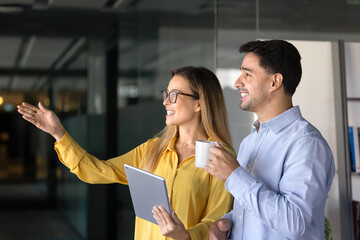 Positive young diverse business colleagues standing in office hall together, looking and pointing hand away, viewing commercial real estate, buildings out of window for investment