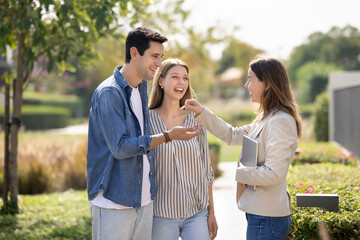 Couple stands outdoors in beautifully landscaped residential complex as cheerful female real estate agent hands over house keys. Moment of successful home purchase, trust between client and realtor © fizkes