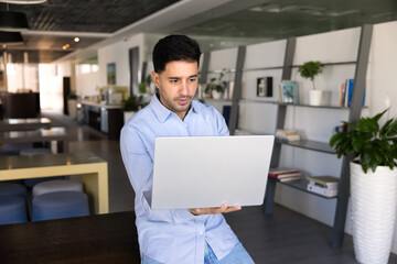Focused young Latin businessman working on urgent task on laptop, standing in co-working space,...