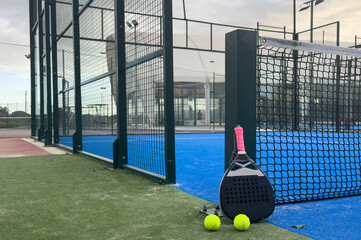 A padel or paddle tennis racket and a bright yellow tennis ball lie on a blue court. The net and a blurred figure of a player are visible in the background.