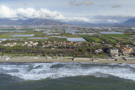 Aerial view of the coastline where the sea's dark waves crash against the sandy shore, contrasting with the bright greenhouses inland, Battipaglia, Campania, Italy.