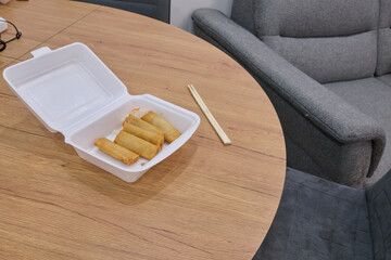Takeout container with crispy spring rolls on a wooden table, accompanied by chopsticks, showcasing a delicious meal experience in a cozy setting